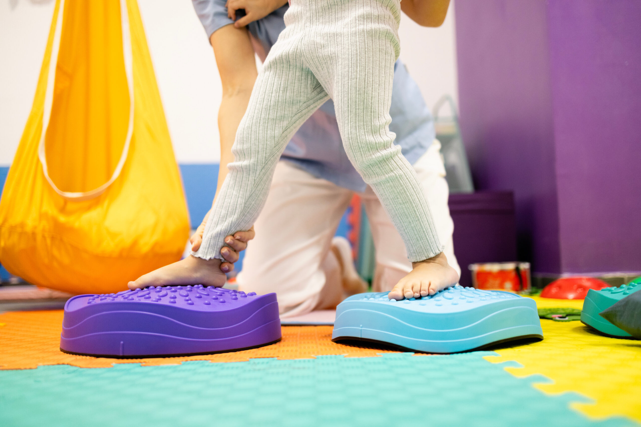 Young child balancing on colorful sensory stepping stones while supported by an adult in a therapy setting